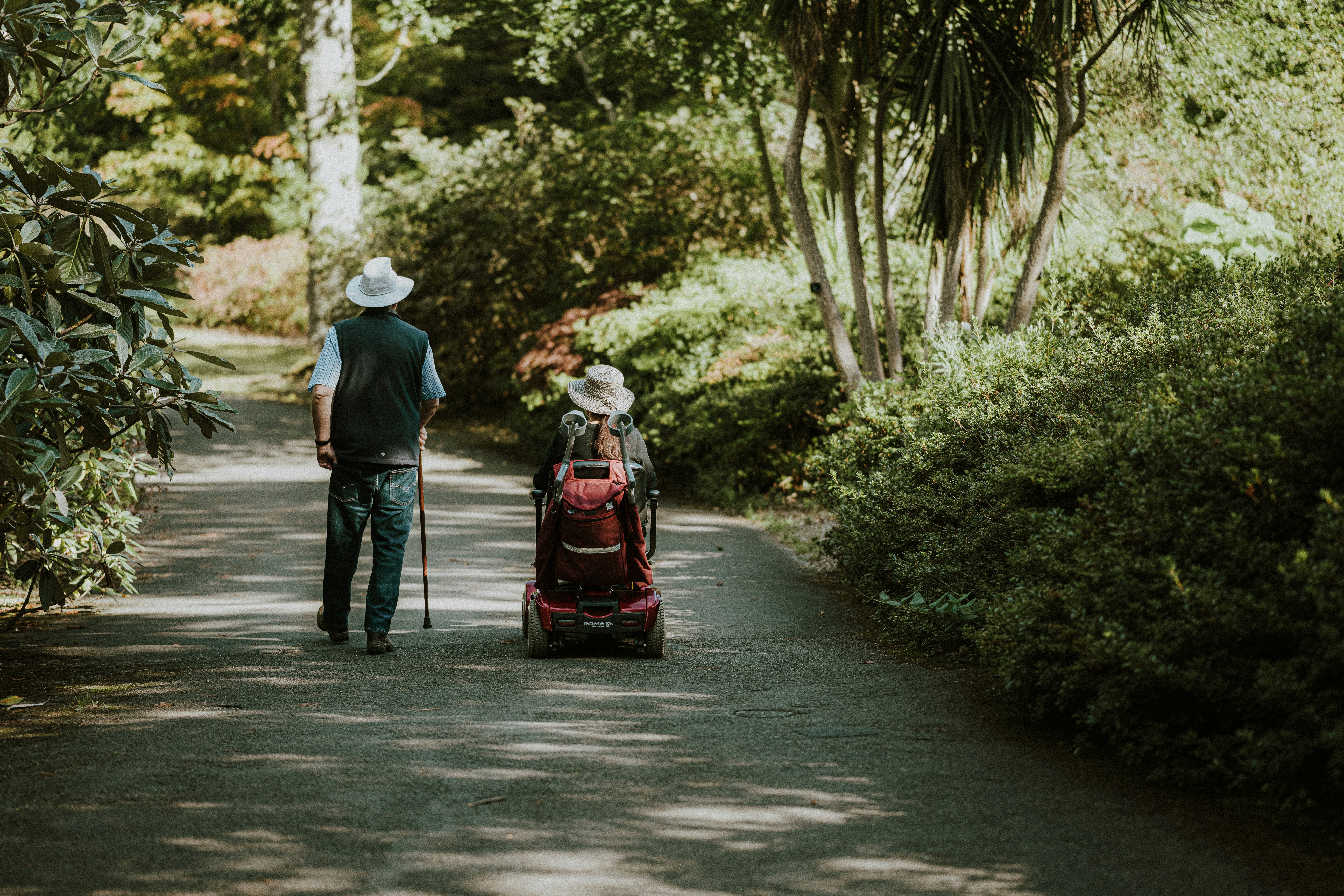 Two people walk side by side along a garden path; one uses a mobility scooter and the other walks alongside.